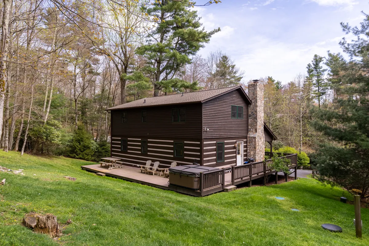 A spacious wooden deck with a stone chimney overlooks a lush green lawn and stands surrounded by tall trees. The deck is set for outdoor dining or gatherings, with ample space for children to play and families to relax after exploring the area. Sunset views and stargazing possibilities are visually implied.