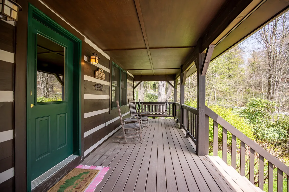 A cozy porch with green doors and classic rocking chairs, bordered by a simple wooden railing and nestled among greenery. The porch faces outward, inviting guests to sit, sip wine, and enjoy the peaceful sounds and sights of the surrounding forest, especially as the sun sets.