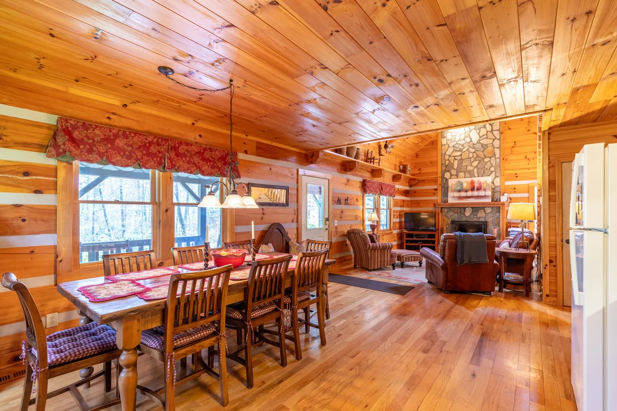 An open-plan interior scene with a spacious dining table, comfortable seating area near the fireplace, and visible exposed wooden beams overhead. The space radiates warmth and is clearly designed for group meals, board games, and shared moments.
