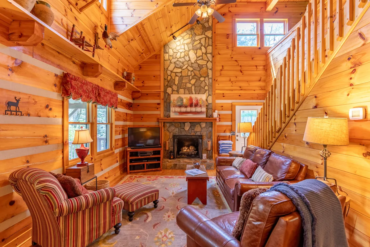 A warmly lit living room featuring a prominent stone gas-log fireplace, plush leather sofa, decorative rug, wooden staircase, and cozy lamps. The soaring ceilings with exposed beams frame the room, creating an inviting gathering space perfect for relaxing or sharing stories by the fire.