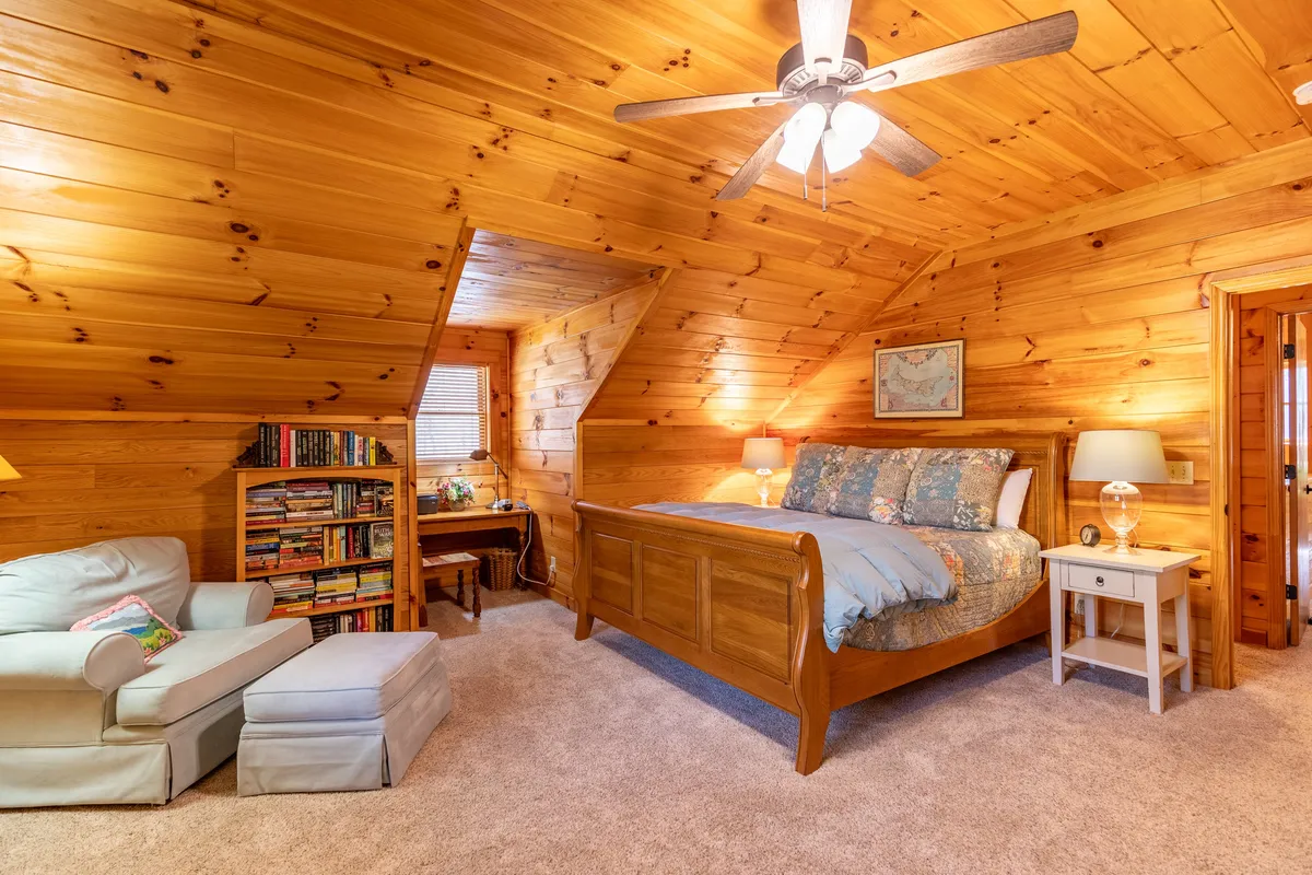 This image features a warm and comfortable bedroom setting with a wooden bed, a cozy armchair, a bookshelf, and a table lamp. Natural light streams in from the window, creating an inviting corner ideal for reading or quiet reflection.
