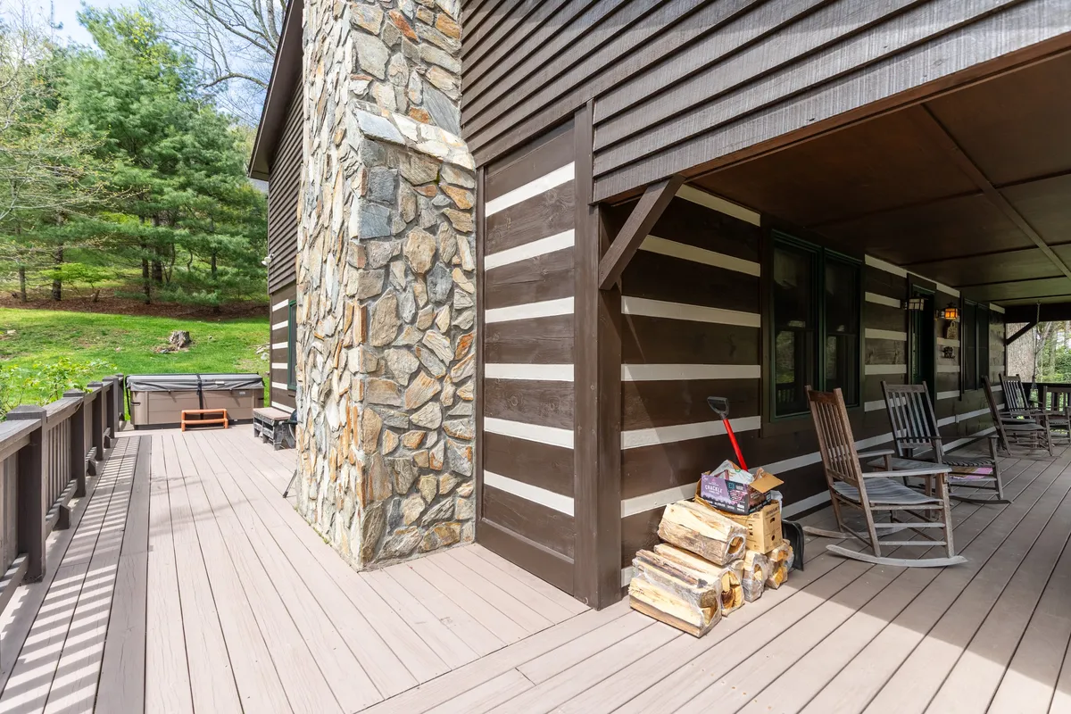 A cozy covered porch featuring classic rocking chairs, stone wall accents, wood decking, and a stack of firewood. The porch is oriented toward scenic greenery, capturing the inviting, peaceful atmosphere ideal for sipping coffee or enjoying a sunset.
