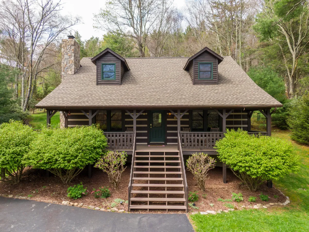 The image captures the inviting front porch of a charming log cabin, complete with cozy rocking chairs and a swing, framed by lush shrubs and a striking stone chimney. The scene suggests a peaceful setting perfect for unwinding, with a green front door adding a welcoming touch and hints of the tranquil natural surroundings.