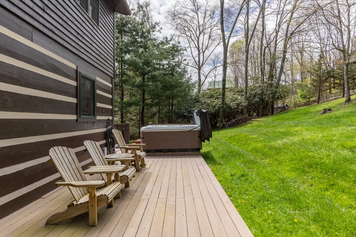This image features a spacious wooden deck surrounded by lush green grass and tall trees. The centerpiece is a hot tub, accompanied by Adirondack chairs that invite relaxation and socialization. The tranquil outdoor setting is ideal for unwinding after a day of outdoor adventures, with sunset views and a peaceful atmosphere.