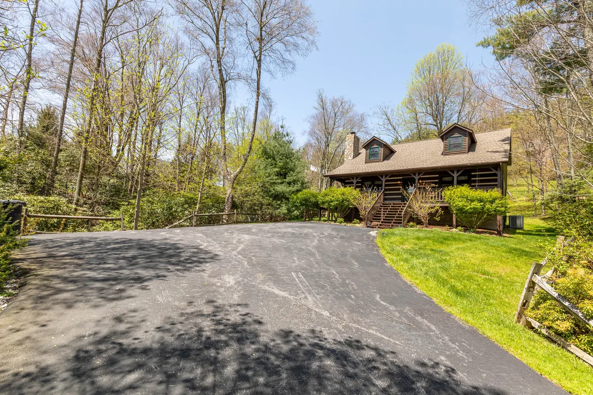 A classic log cabin sits at the end of a winding driveway, nestled among lush greenery. The porch offers inviting views, perfect for morning coffee or watching the sunset after a day of adventure. The natural setting immediately shows the peaceful, family-friendly environment and proximity to local attractions.