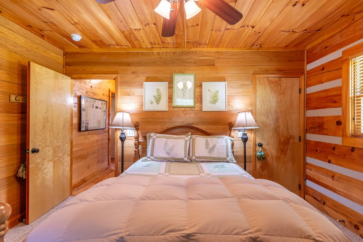 A cozy bedroom featuring a comfortable bed with plush bedding, warm wooden walls, ambient lamps, and decorative prints on the wall. The space is filled with soft natural light, creating an intimate and serene retreat for relaxation or restful sleep.