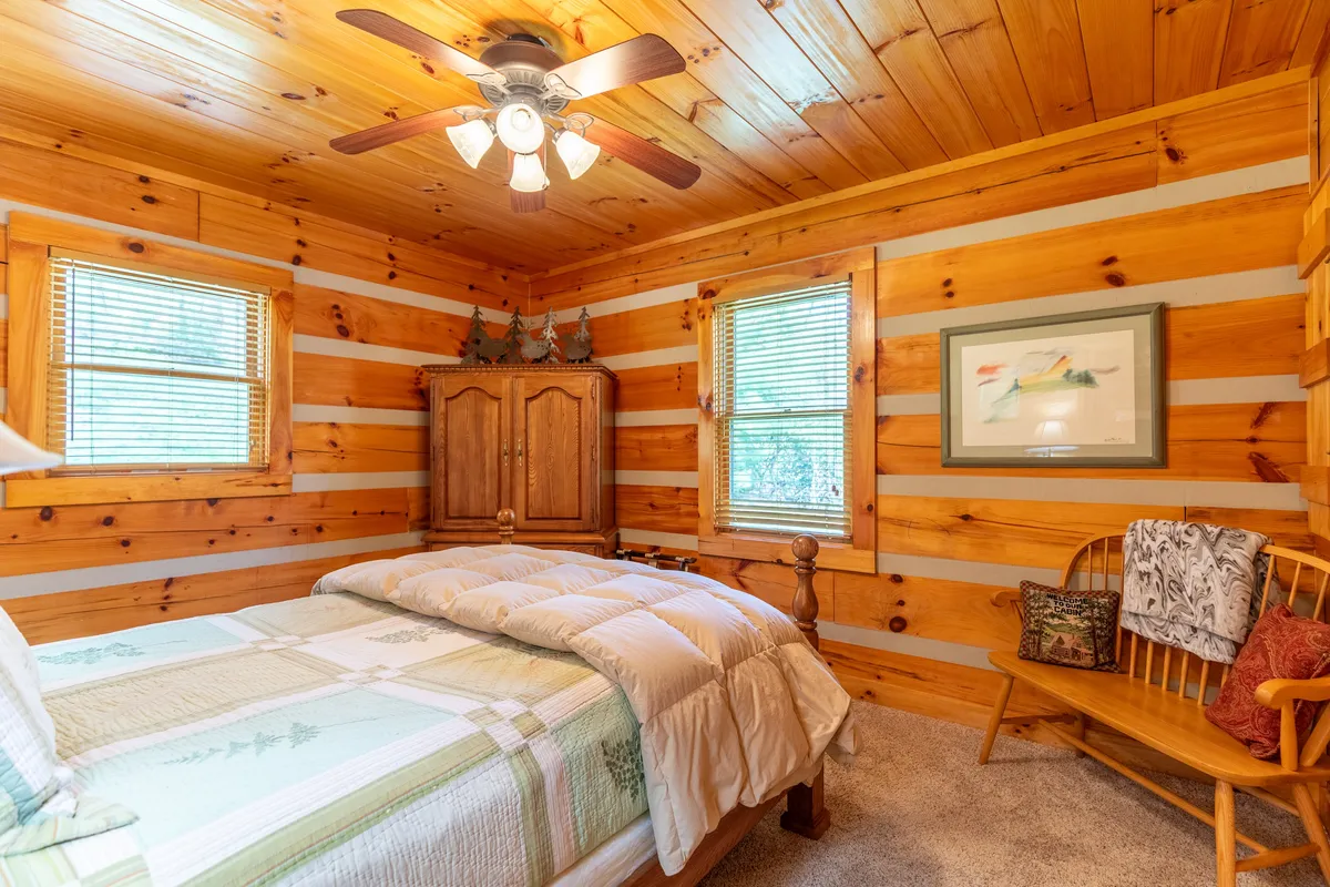 A spacious bedroom with wood paneling, a comfortable bed topped with inviting linens, an armoire for storage, and windows that allow in plentiful daylight. The room feels open yet cozy, ideal for a restful night or leisurely morning.